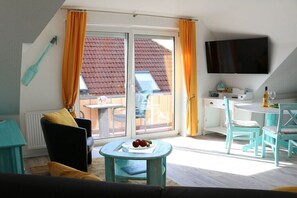 Ein Zimmer mit einer Couch, einem Tisch mit Früchten, einem Fernseher und einem Balkon mit orangenen Vorhängen.
