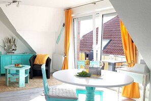 Ein Essbereich mit einem runden Tisch, Stühlen, einem kleinen Beistelltisch und Blick ins Freie durch ein Fenster mit Vorhängen.