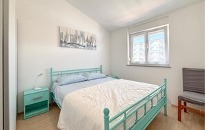 Ein Schlafzimmer mit Bett, Nachttisch, Stuhl, einem Fenster mit floralem Rollo und einem Stadtbild an der Wand.