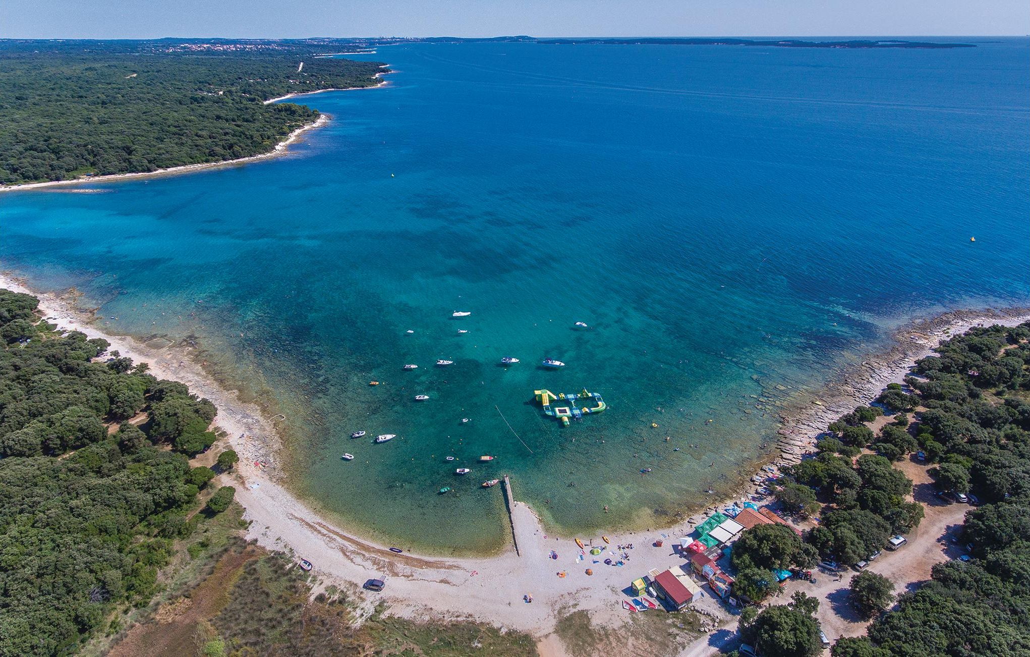 Luftaufnahme einer Küstenlandschaft mit klarem Wasser, Booten und einem Strand mit Bauten und Grünflächen.