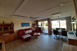 Ein Wohnzimmer mit rotem Sofa, Holzschrank, Esstisch mit Stühlen und Blick durch die Glastür.