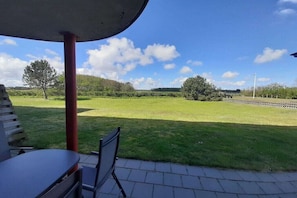 Eine Terrasse im Freien mit Tisch und Stühlen, Blick auf ein grünes Feld und einen klaren Himmel.