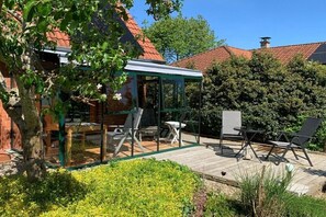 Eine überdachte Terrasse mit Gartenmöbeln, einer Glaseinfriedung und einem Backsteinhaus mit rotem Ziegeldach.