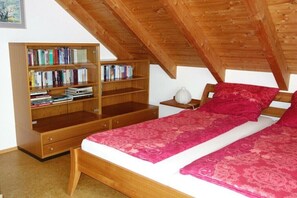 Ein Schlafzimmer mit Holzboden, einem Bett mit roter Bettdecke, einem Nachttisch mit Lampe und einem Bücherregal voller Bücher.