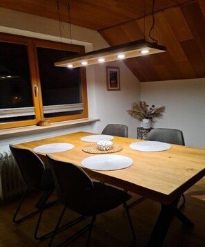 Ein Essbereich mit einem Holztisch, schwarzen Stühlen, einer Pendelleuchte, einem Fenster mit Jalousien und einer Vase mit Trockenpflanzen.
