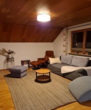 Ein gemütliches Wohnzimmer mit Holzbalkendecke, einem grauen Sofa, einem braunen Ledersessel, einem kleinen runden Tisch und einem Fenster mit Vorhängen.