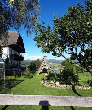 A wooden play structure with a slide, surrounded by a grassy area with a house and trees in the background. - Organic farm with 1 bedroom (Mariapfarr)