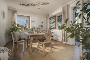Ein Essbereich mit Holztisch und Stühlen, einer Kandelaberleuchte und großen Fenstern mit Blick auf die Berge.