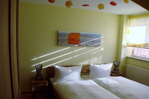 Ein Zimmer mit einem Bett, einem kleinen Tisch, einer Lampe, einem Bild an der Wand und einem Fenster mit Vorhängen.