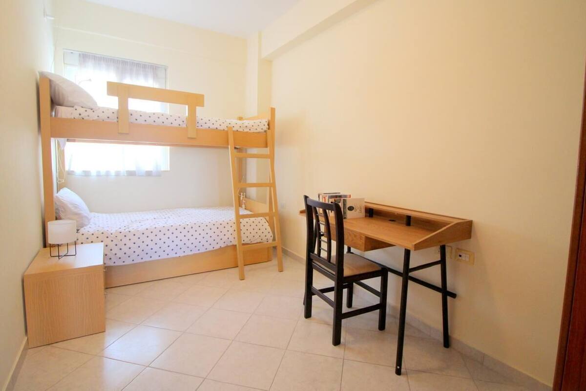 A bunk bed with a desk and chair, a bedside table, and a window with curtains.