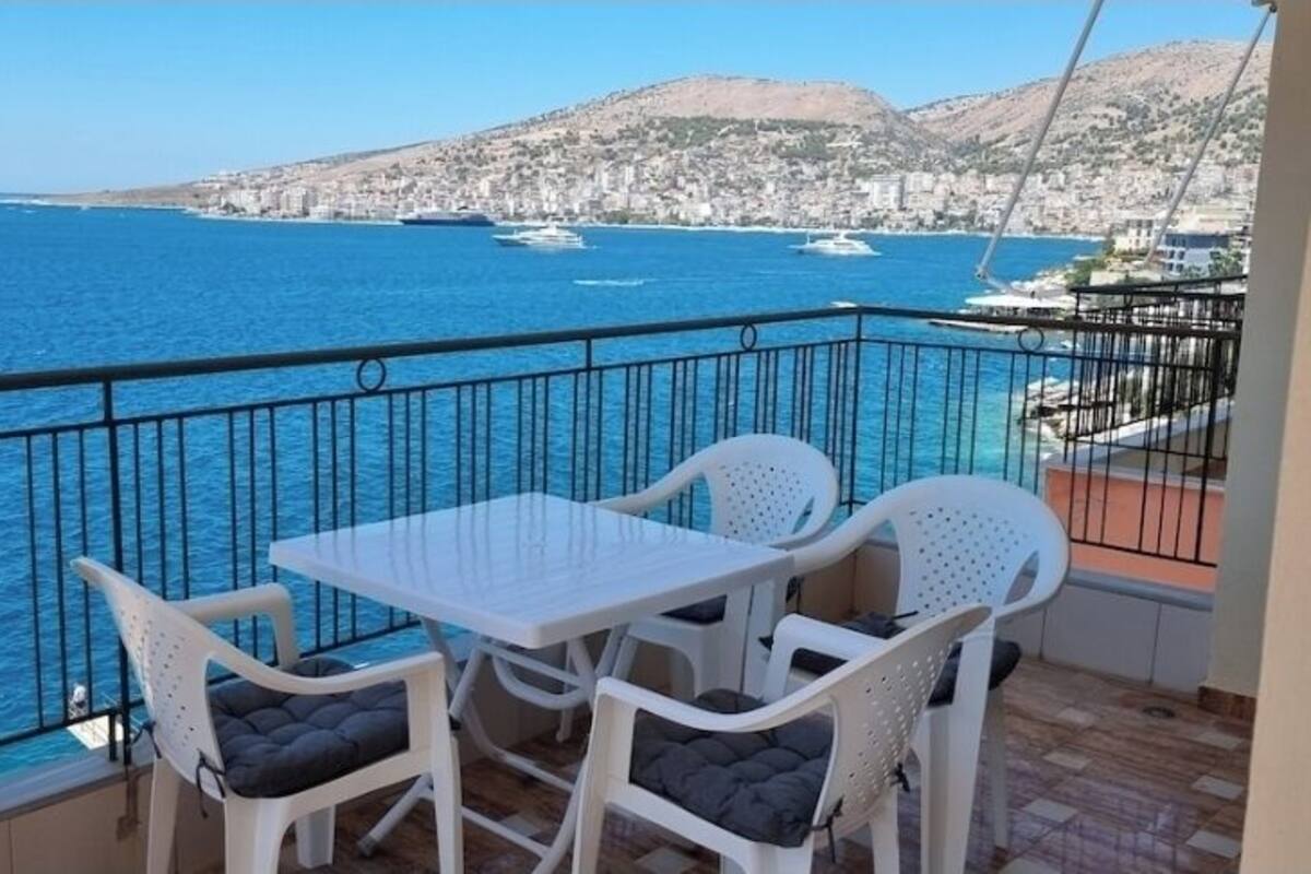 A balcony with a table and chairs, overlooking a coastal city with a clear view of the sea and boats.