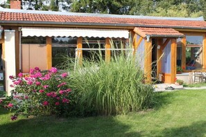 Ein Haus mit Ziegeldach, einer überdachten Veranda und einem Garten mit rosa Blumen und hohem Gras.