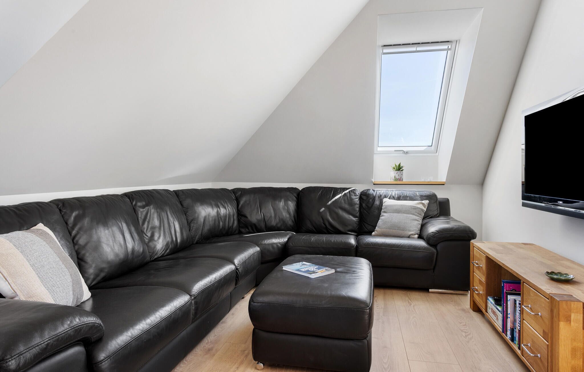 Uma sala de estar moderna com um sofá seccional, um pequeno banquinho, um aparador de madeira e uma televisão de tela plana.