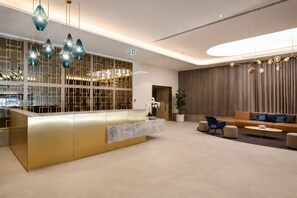 Ein moderner Empfangsbereich mit einer goldenen Theke, blauen Pendelleuchten, einer Sitzecke mit Sofa und Sesseln sowie einer großen Pflanze in einem Topf.