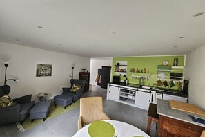 Ein modernes Wohnzimmer mit einer Sofagarnitur, einem Esstisch mit Stühlen, einer Küchenzeile mit grüner Rückwand und einem Bücherregal.