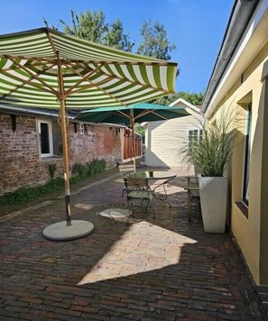 Ein Innenhof mit Backsteinpflaster, einem grün-weiß gestreiften Sonnenschirm, einem Tisch mit Stühlen und einem Blumentopf.