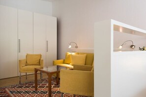 Ein Zimmer mit einer Couch, einem Sessel und einem kleinen Tisch. Zwei Wandlampen und ein Kleiderschrank sind ebenfalls vorhanden.