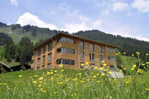 Ein mehrstöckiges Gebäude mit Holzfassade, eingebettet in eine gebirgige Landschaft und ein Feld mit gelben Blumen.