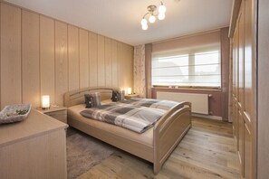Ein Schlafzimmer mit einem Bett, Nachttischen, einem Fenster mit Jalousien, einem Heizkörper und einem Holzschrank.