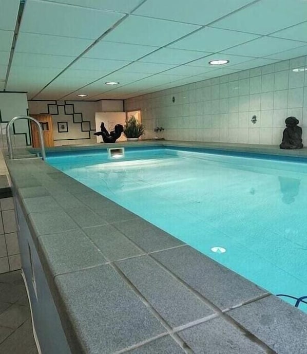 Ein Schwimmbecken mit einer Fliesenveranda, einer Metallleiter und einer Statue auf der rechten Seite.