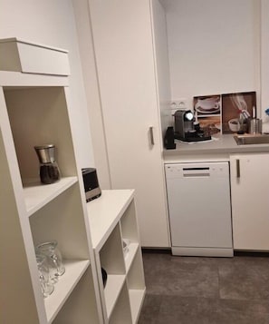 Eine kompakte Küche mit weißen Schränken, einer Kaffeemaschine, einem Geschirrspüler und einer kleinen Arbeitsplatte mit Spüle.
