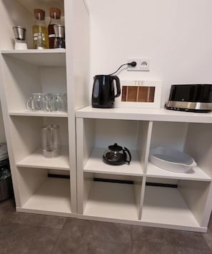 Ein weißes Regal mit verschiedenen Küchenutensilien, darunter eine schwarze Kaffeemaschine, eine Teekanne aus Glas und eine Teeauslage.