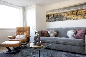 Ein Wohnzimmer mit einem Ledersessel, einem grauen Sofa, einem Glastisch und einem Gemälde, das eine Strandansicht zeigt.