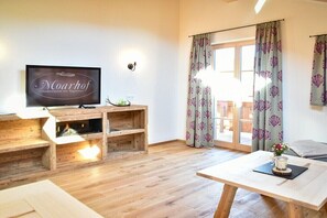 Ein gemütliches Wohnzimmer mit Fernseher, einer Holzunterhaltungsecke und Blick ins Freie durch eine Glastür.