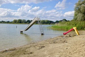 A beach with a metal slide partially submerged in water, a ladder, and a red and yellow play structure. - Cozy thatched cottage (Klocksdorf)
