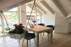 Ein Essbereich mit einem Holztisch und -stühlen, einem hängenden Korb mit Pflanzen und großen Fenstern mit Blick auf Grünflächen.