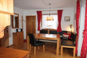 Ein Zimmer mit einem Holzesstisch, schwarzen Stühlen, einer Couch mit roten Vorhängen, einem Holzkasten und einem Spiegel.