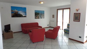 Ein Wohnzimmer mit einer roten Sofagarnitur, einem Fernseher auf einem Schrank und zwei großformatigen Landschaftsbildern an den Wänden.