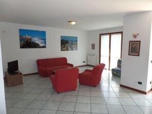 Ein Wohnzimmer mit einer roten Sofagarnitur, einem Fernseher auf einem Schrank und zwei großformatigen Landschaftsbildern an den Wänden.