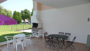 Eine Terrasse im Freien mit Grill, weißen Kunststoffstühlen und einem grünen Tisch.