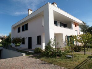 Ein modernes, zweistöckiges Haus mit flachem Dach, großen Fenstern und einem kleinen Garten mit Sträuchern und einem Baum.