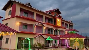 A multi-story hotel with a red roof, balconies, and a well-lit outdoor area with seating. - Dev murti eco resort  (Mal)