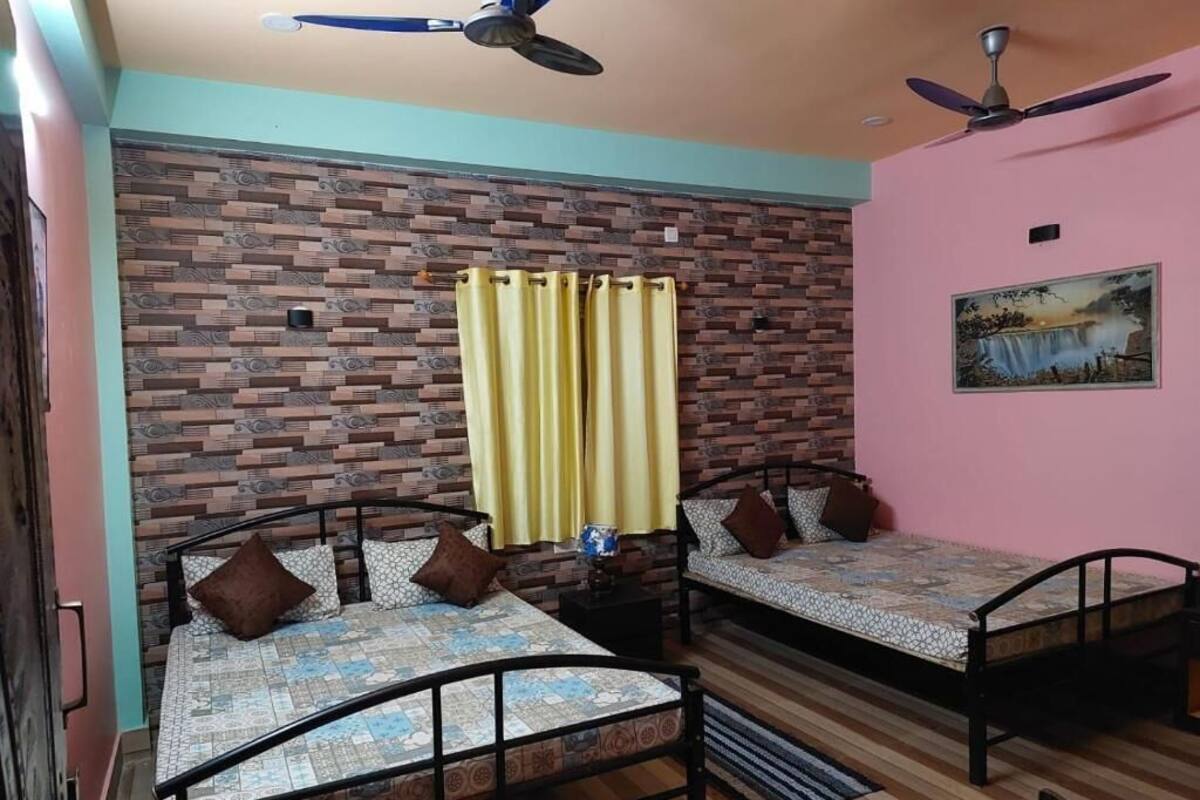 A room with two beds, a brick-patterned wall, a ceiling fan, a painting, and a window with yellow curtains.