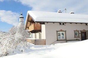 Ein schneebedecktes Gebäude mit einem hölzernen Balkon, einem Fenster mit dekorativem Rahmen und einem im Hintergrund sichtbaren Glockenturm.