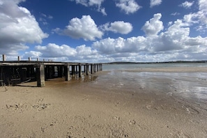 Ein Strand mit einer Holzsteg, der sich in ruhiges Wasser erstreckt, Sandstrand und teils bewölkter Himmel.