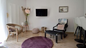 Ein Wohnzimmer mit Fernseher, einem Gemälde, einem Korbstuhl, einem schwarzen Sessel, einem violetten Teppich und einem kleinen runden Tisch.
