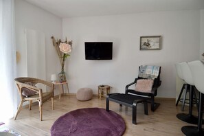 Ein Wohnzimmer mit Fernseher, einem Gemälde, einem Korbstuhl, einem schwarzen Sessel, einem violetten Teppich und einem kleinen runden Tisch.