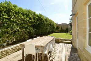 Eine Holzterrasse mit Tisch und Stühlen, eine Steinmauer und ein Heckenstreifen.