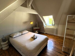 Ein Schlafzimmer mit schrägem Dach, einem Bett mit Kissen, einem Nachttisch mit Lampe, einem Heizkörper und einem Fenster mit Tageslicht.