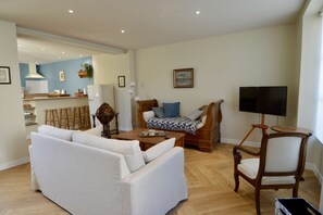 Ein Wohnzimmer mit einer Couch, einem Holzstuhl, einem Couchtisch, einem Fernseher auf einem Schrank und einer Küche im Hintergrund.