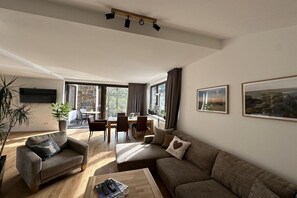 Ein Wohnzimmer mit Sofa, Sessel, Couchtisch und Fernseher. Es gibt eine Essecke mit Stühlen und einem Tisch.