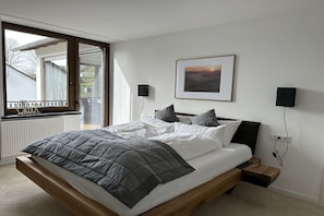 Ein modernes Schlafzimmer mit einem großen Bett, einem Fenster mit Ausblick, einem gerahmten Bild an der Wand und einem Nachttisch mit Lampe.