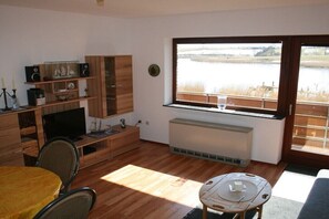 A modern living room with a TV, wooden furniture, a dining table, and a large window overlooking a body of water. - Water chicks at the boat dock (Kosel-Missunde)