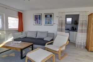 Ein modernes Wohnzimmer mit einer Couch, Sesseln und einem Couchtisch, sowie einer Küche im Hintergrund und zwei gerahmten Kunstwerken an der Wand.