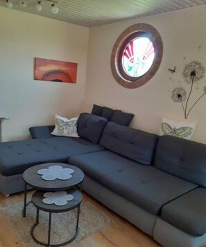 Ein modernes Wohnzimmer mit einer Eckcouch, zwei kleinen runden Beistelltischen, einem Rundfenster und Wandbildern.