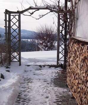 Ein hölzerner Bogen mit einem mit Schnee bedeckten Weg, der darauf führt, einem Holzstapel und einem Gebäude im Hintergrund.